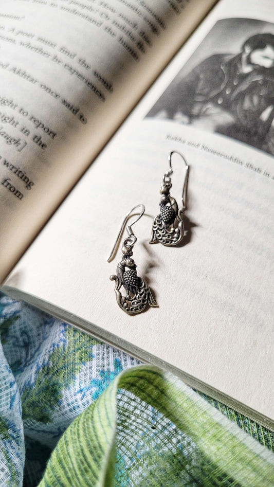 Pair of Mayil silver earrings on an open book with a green fabric background