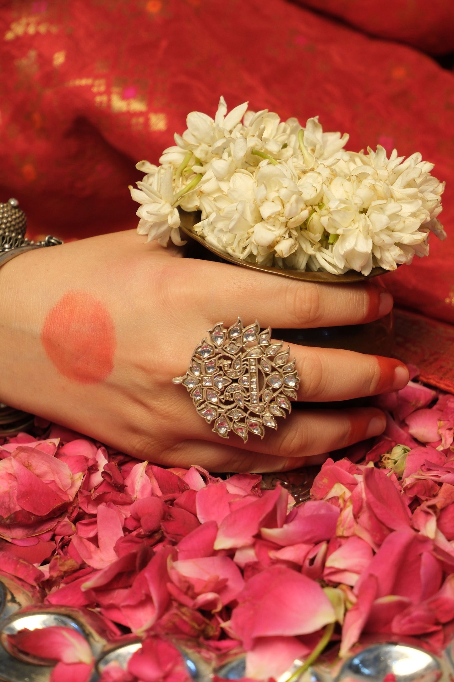 Close-up of a hand with a decorative ring, surrounded by pink flowers and a red fabric background.