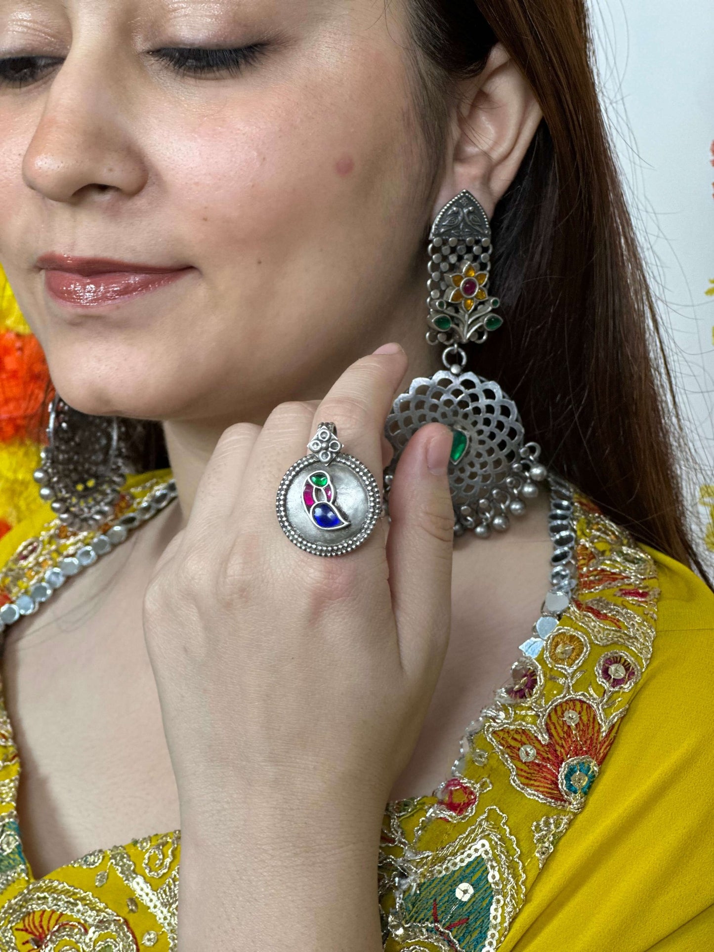 Woman wearing silver earrings and a ring with a colorful design, against a blurred background.