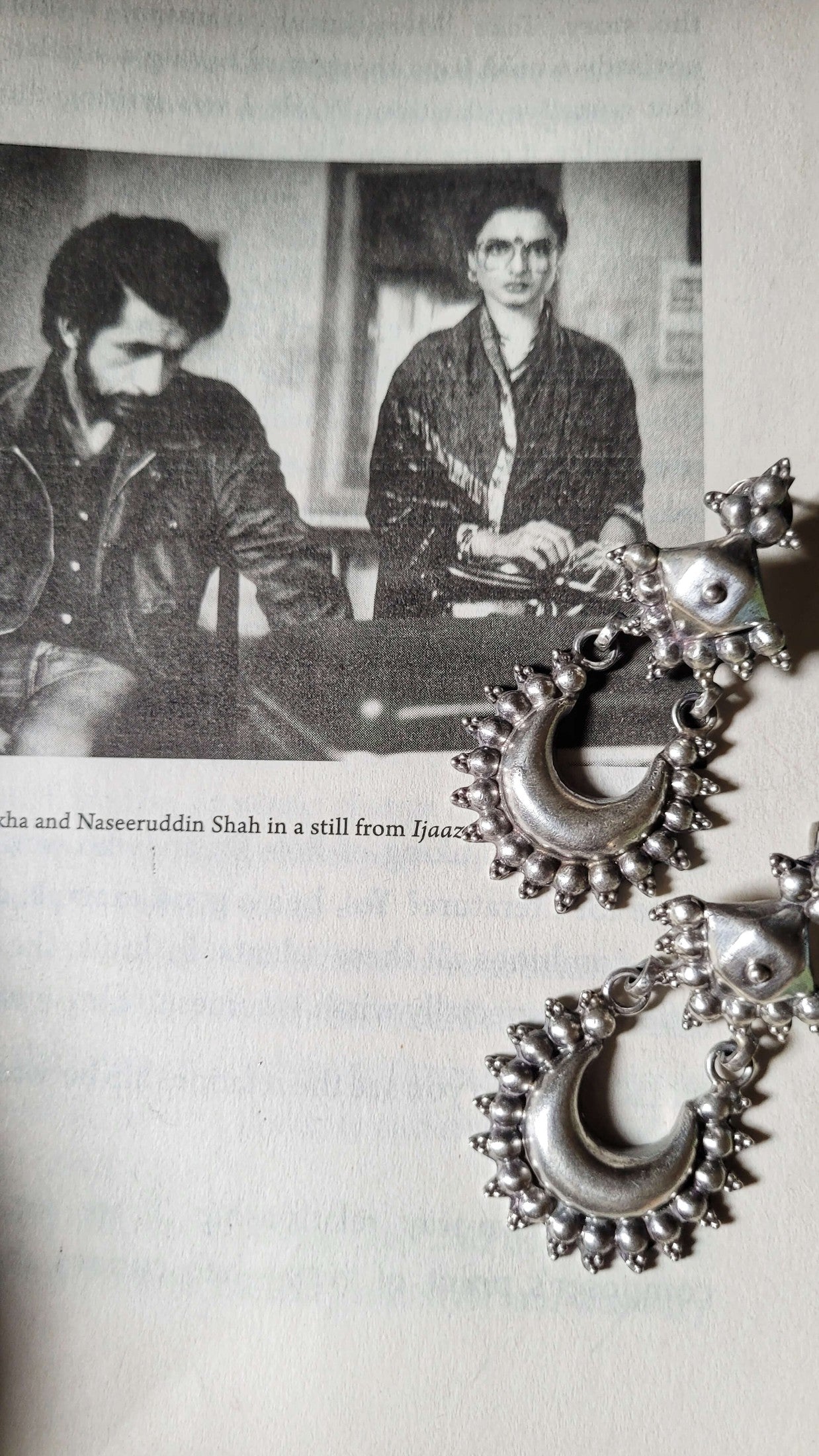 Silver earrings with crescent moon design on a light background, with a black and white photo of two people in the background.