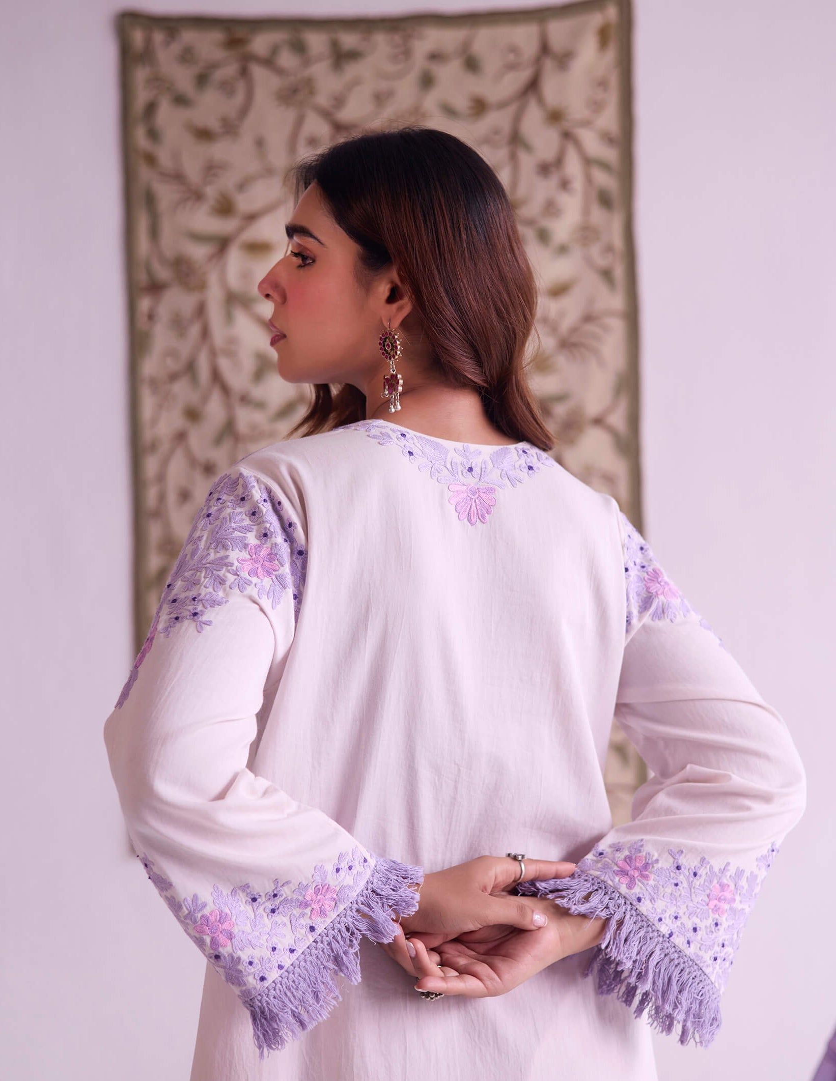 Woman wearing a white outfit with purple embroidery in a room with a decorative wall hanging.