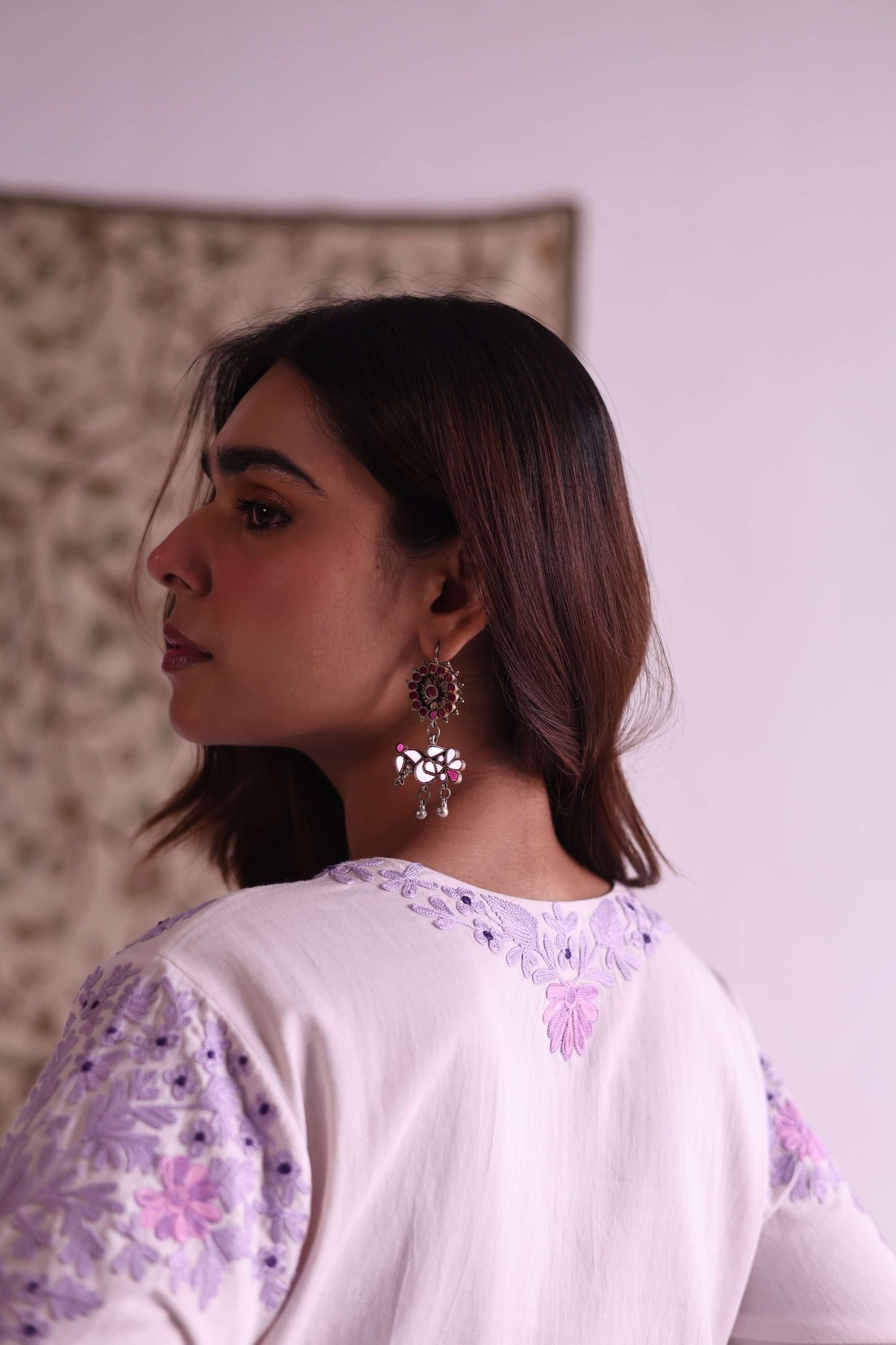 Woman wearing a white top with floral patterns, standing against a plain wall.