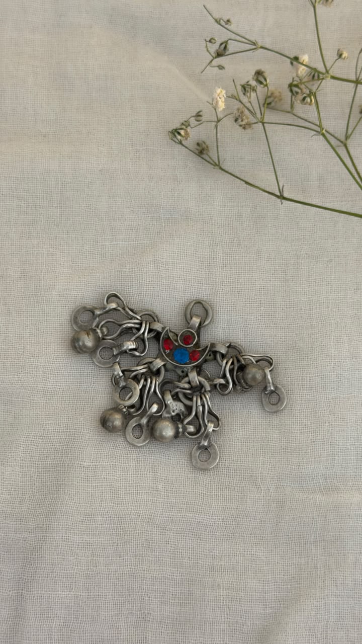 A silver-toned metal decorative piece with multiple dangling loops and a small red and blue center detail, placed on a light beige fabric background with dried flowers in the upper right corner.
