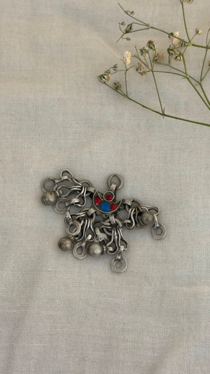 A silver-toned metal decorative piece with multiple dangling loops and a small red and blue center detail, placed on a light beige fabric background with dried flowers in the upper right corner.