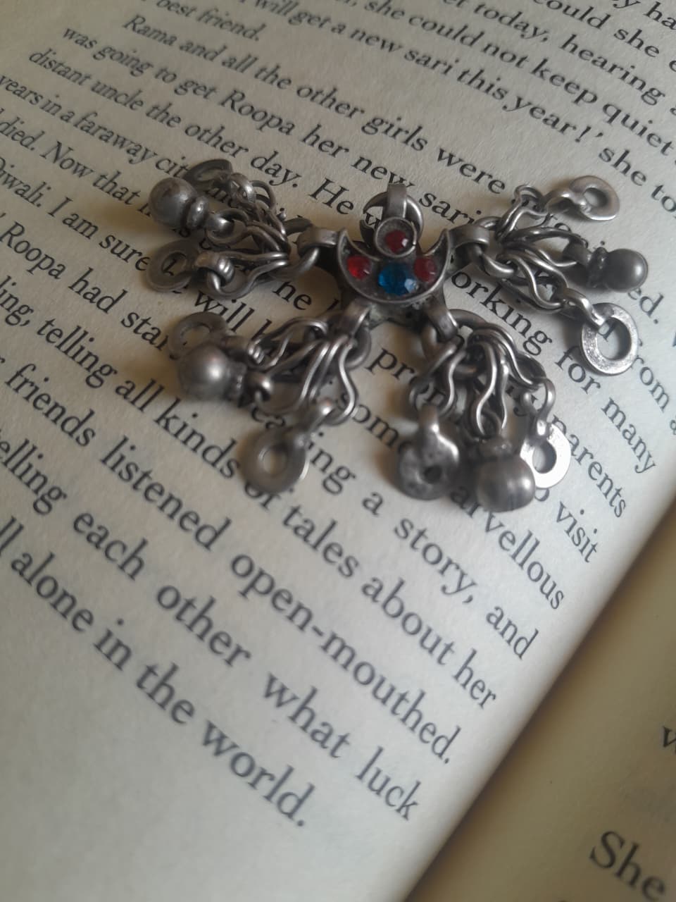 A silver-toned metal cross pendant with dangling charms and a small red and blue stone in the center, resting on an open book with printed text.