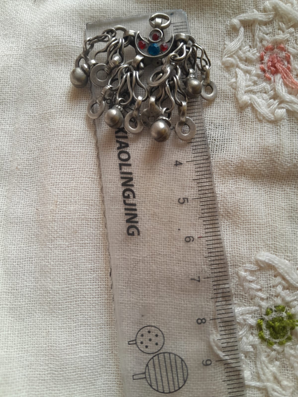 A silver-toned metal charm accessory with multiple dangling loops and a small red-and-blue decorative center, placed on top of a clear plastic ruler over a cream fabric background.