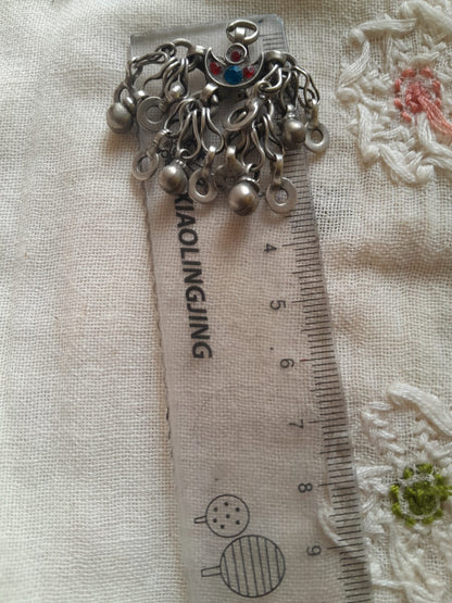 A silver-toned metal charm accessory with multiple dangling loops and a small red-and-blue decorative center, placed on top of a clear plastic ruler over a cream fabric background.