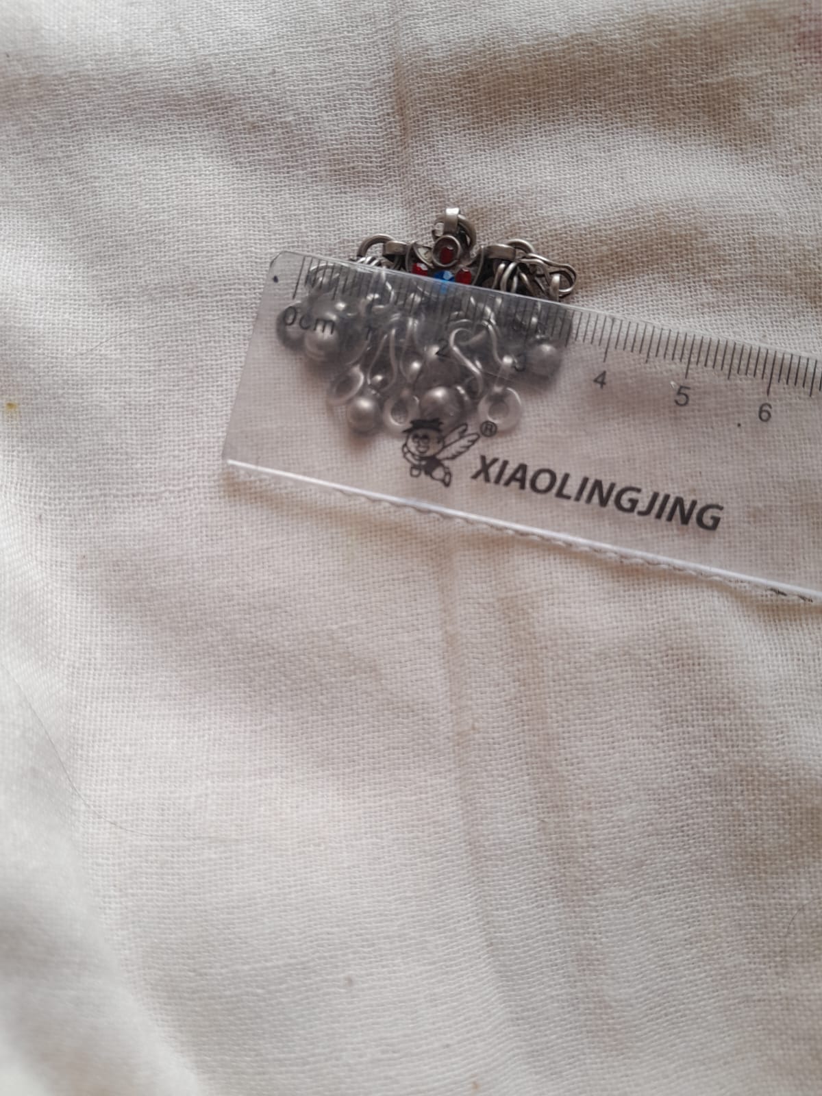 A small cluster of silver-toned metal beads or charms with a ring attached, placed on a clear plastic ruler over a light fabric background. 