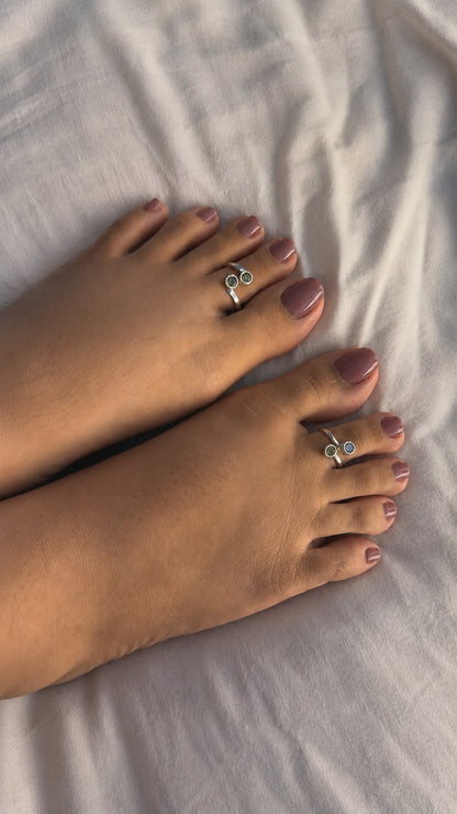 A close-up of a person's feet resting on white fabric, showcasing two silver toe rings with small clear stones set on the big toes.
