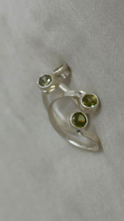 A silver-toned ring with three oval green gemstones in bezel settings, displayed on a textured light gray fabric background.