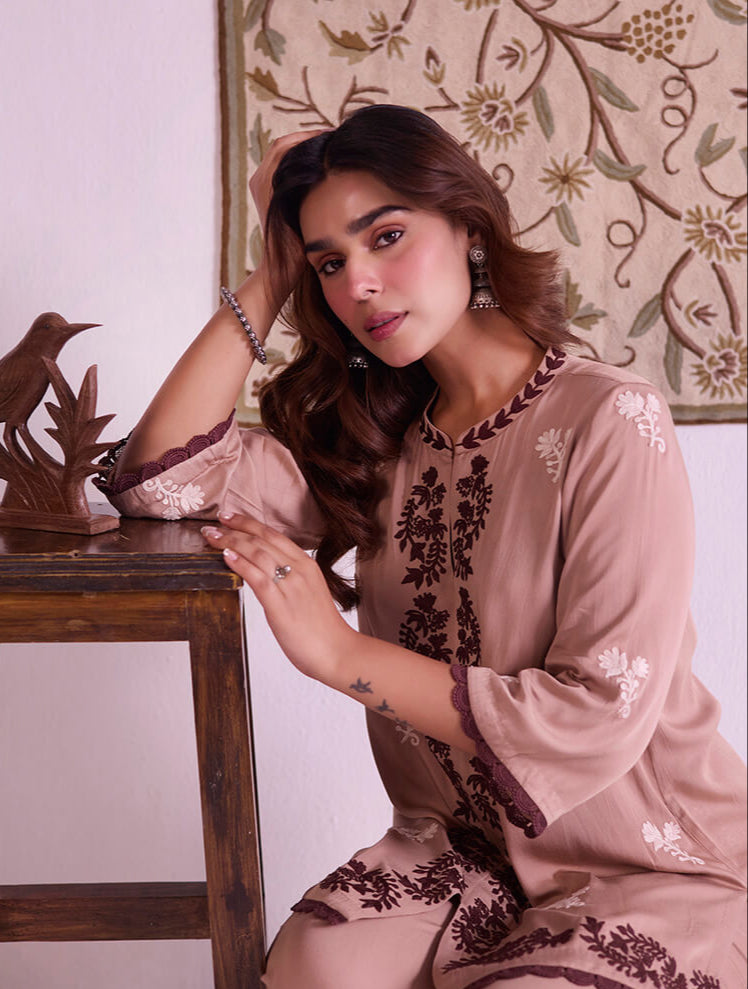 Woman in a beige and brown embroidered outfit sitting on a wooden stool.