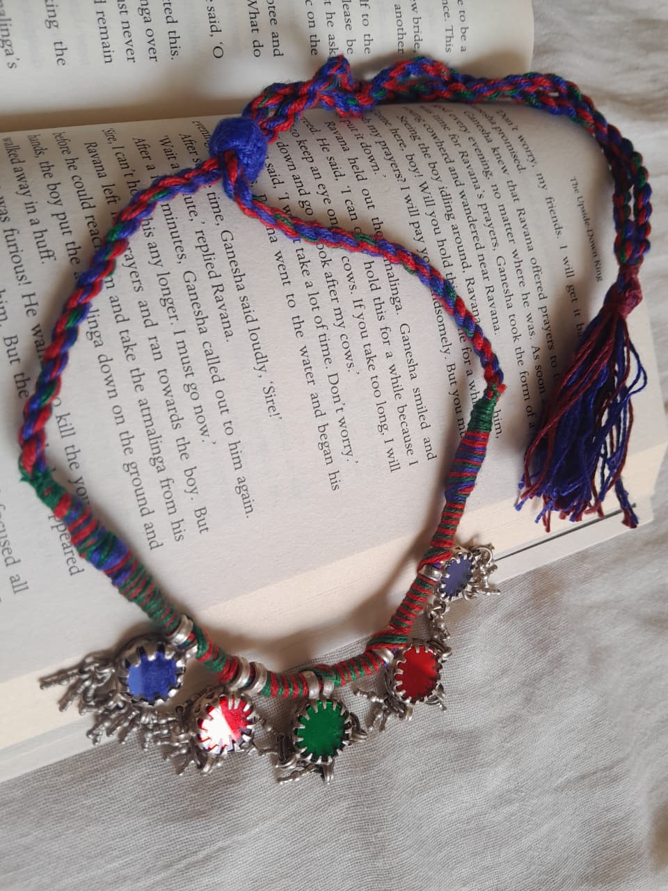 A colorful braided necklace with silver-toned star-shaped charms and blue, red, green, and white stones, laid on top of an open book.