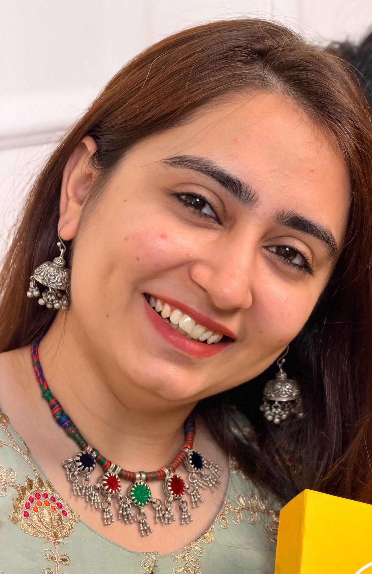 A smiling woman with brown hair wearing a colorful beaded necklace with silver-toned jhumka-style charms and matching earrings, holding a bright yellow box in a close-up indoor setting.