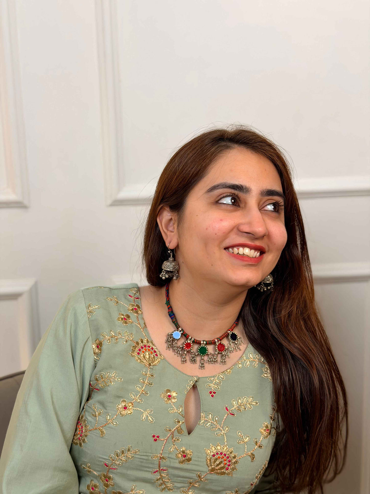 A woman with long brown hair is shown from the chest up, wearing a sage green embroidered top with gold and red floral patterns, a colorful beaded necklace, and silver earrings. She is looking to the side against a white paneled wall background.