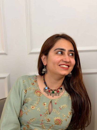 A woman with long brown hair is shown from the chest up, wearing a sage green embroidered top with gold and red floral patterns, a colorful beaded necklace, and silver earrings. She is looking to the side against a white paneled wall background.