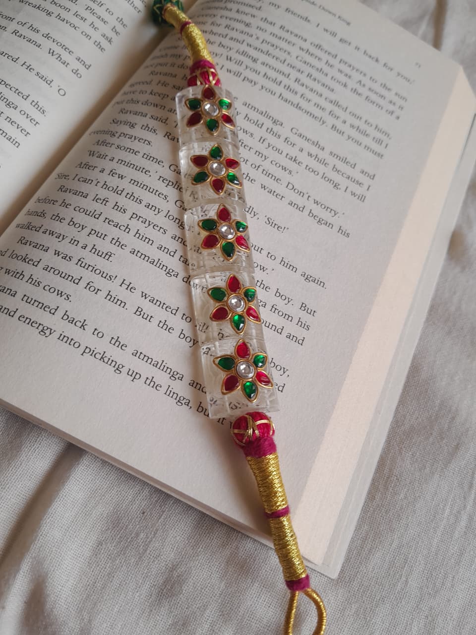 A handmade Indian-style beaded wrist chain (paunchi) with a clear quartz central section, red and green stone floral motifs, and gold-plated wire wrapping, resting on an open book.