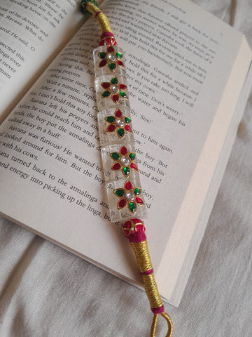 A handmade Indian-style beaded wrist chain (paunchi) with a clear quartz central section, red and green stone floral motifs, and gold-plated wire wrapping, resting on an open book.