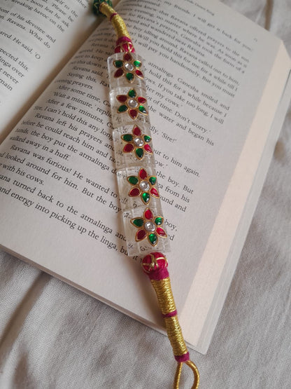 A handmade Indian-style beaded wrist chain (paunchi) with a clear quartz central section, red and green stone floral motifs, and gold-plated wire wrapping, resting on an open book.