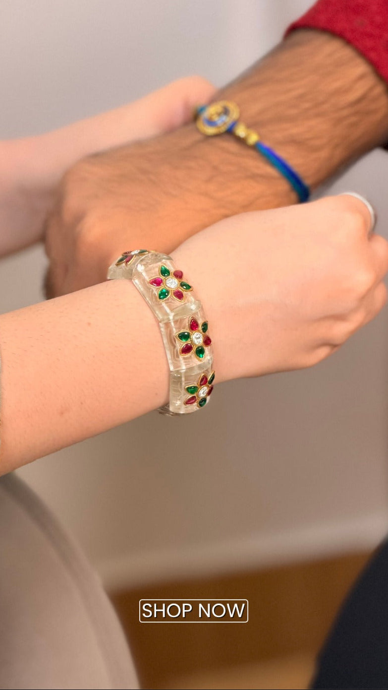 A close-up lifestyle shot of a person wearing a clear glass bangle bracelet decorated with red and green stones. Another person's hand is resting on their shoulder, wearing a blue and yellow beaded bracelet.