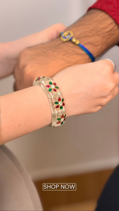 A close-up lifestyle shot of a person wearing a clear glass bangle bracelet decorated with red and green stones. Another person's hand is resting on their shoulder, wearing a blue and yellow beaded bracelet.