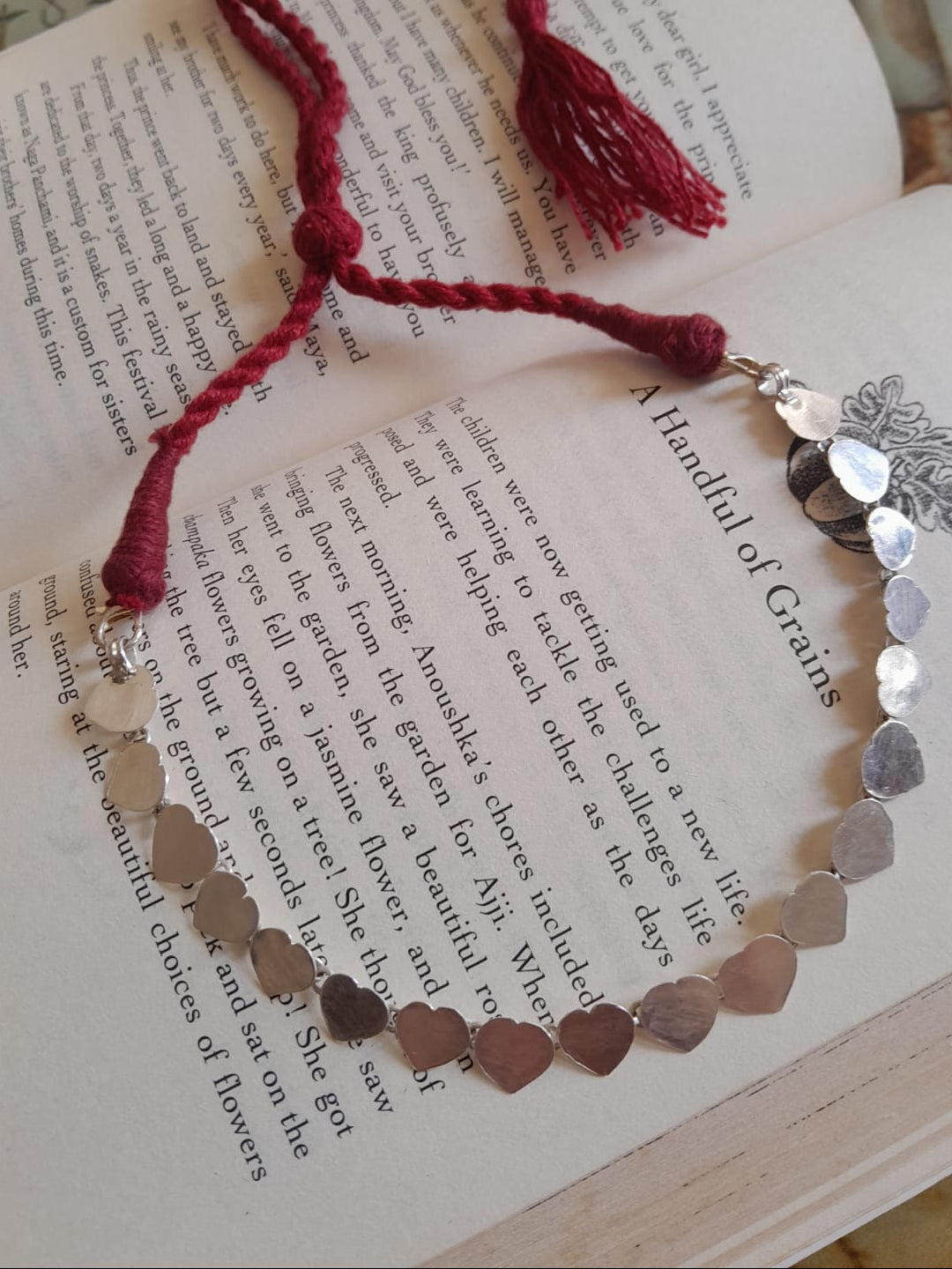 A handmade pendant necklace made of silver heart-shaped charms connected by a red cord with tassel ends, displayed on an open book.