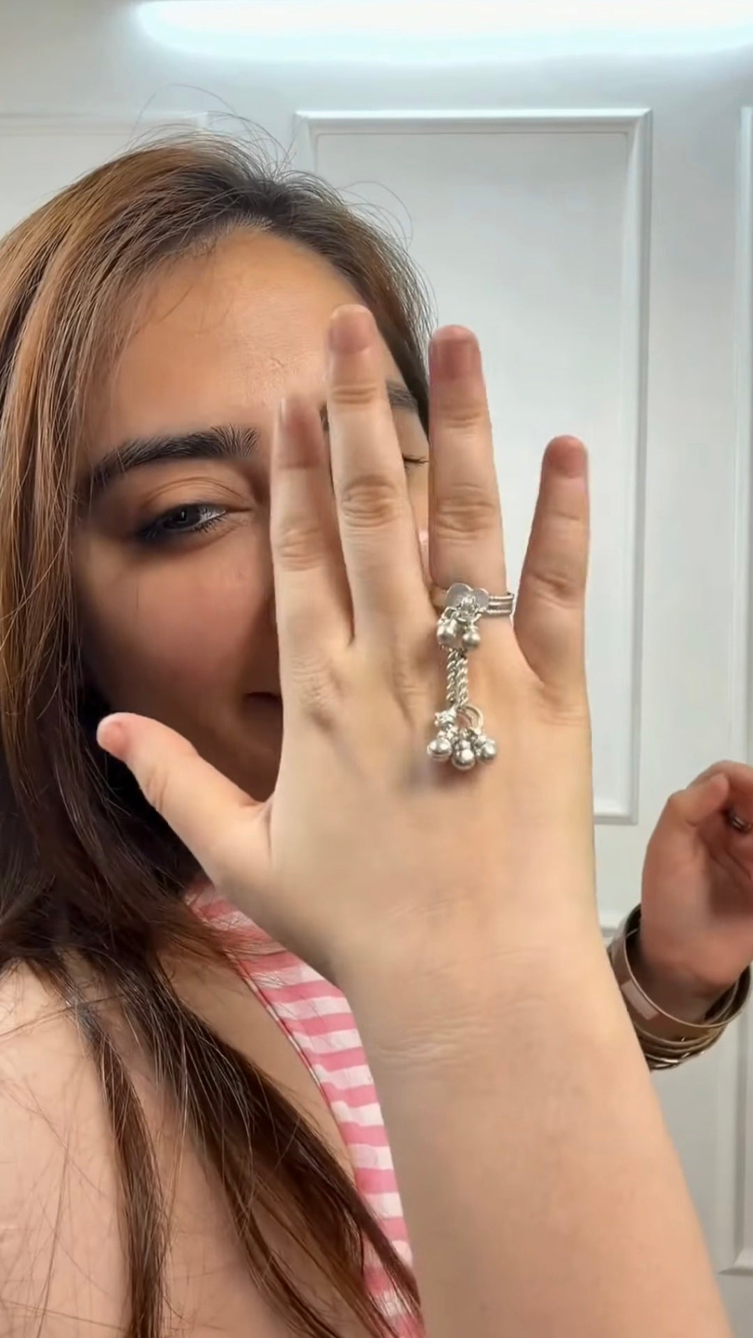 A woman holding her hand up to show a silver-toned key-shaped ring with ghungroos on her finger, set against a white paneled door background.