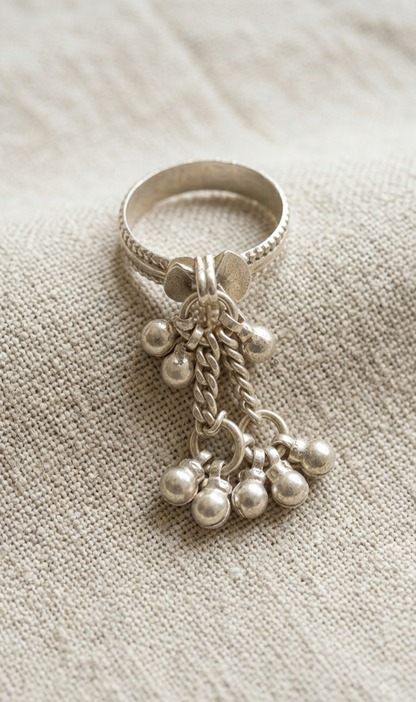 A silver-toned ring with a textured band and dangling charm elements, including small bead clusters and a twisted rope detail, photographed on a neutral beige fabric background.