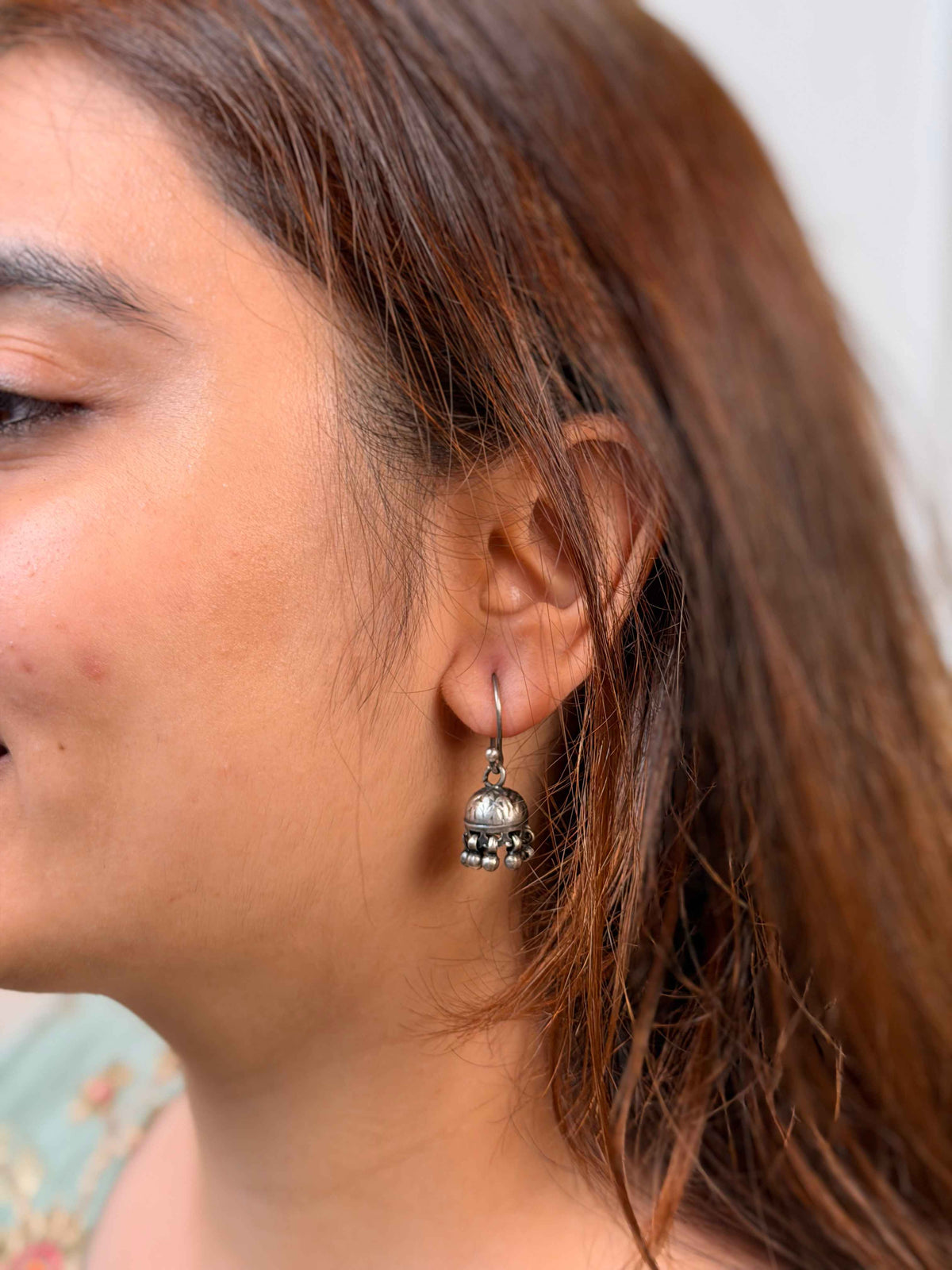 A close-up side profile of a woman wearing a small silver-toned dangle earring with a circular charm and dangling bead details.