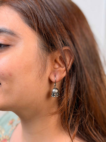 A close-up side profile of a woman wearing a small silver-toned dangle earring with a circular charm and dangling bead details.