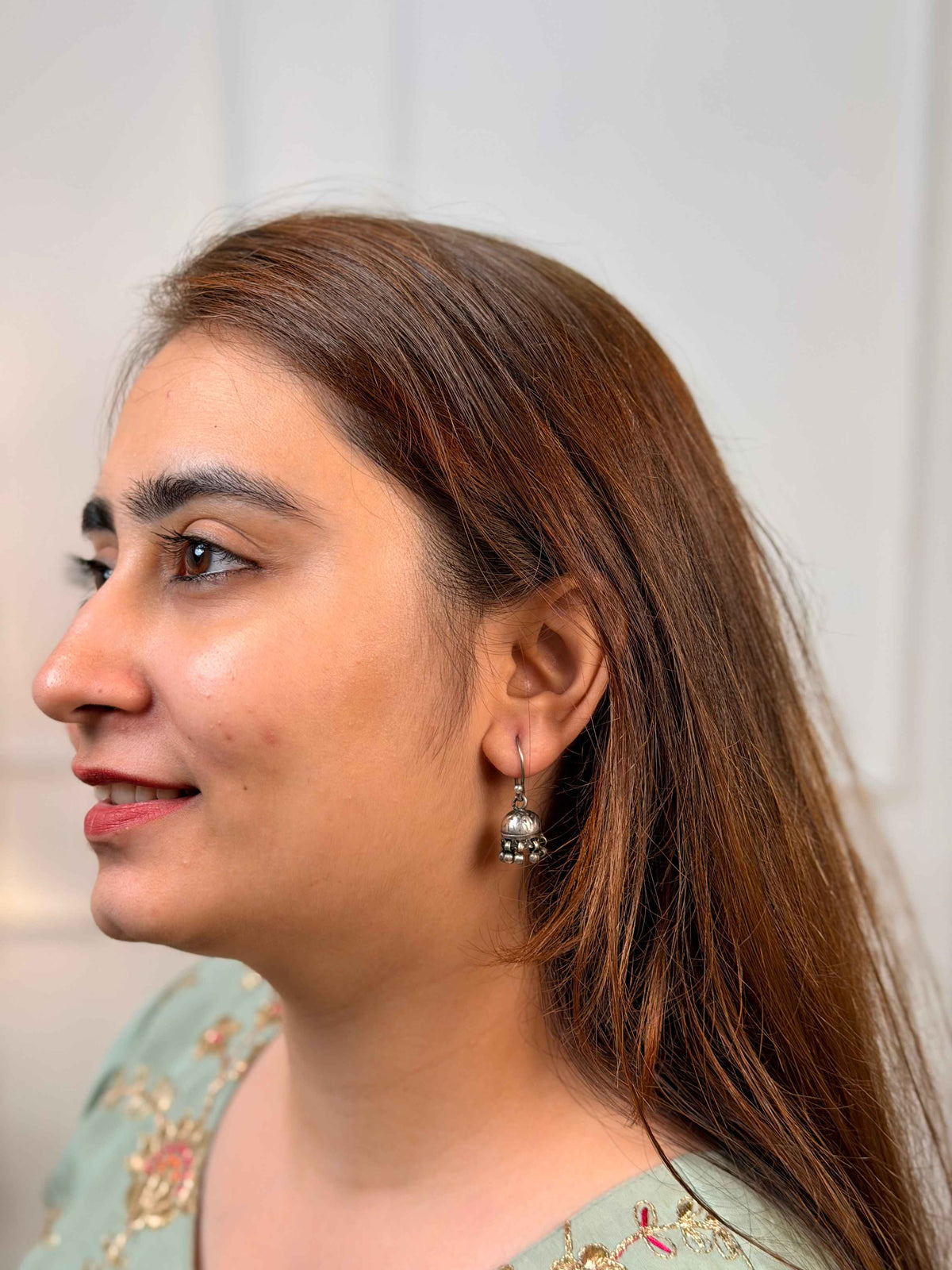 A close-up side profile of a woman wearing a small silver-toned dangle earring with a circular charm and dangling bead details.