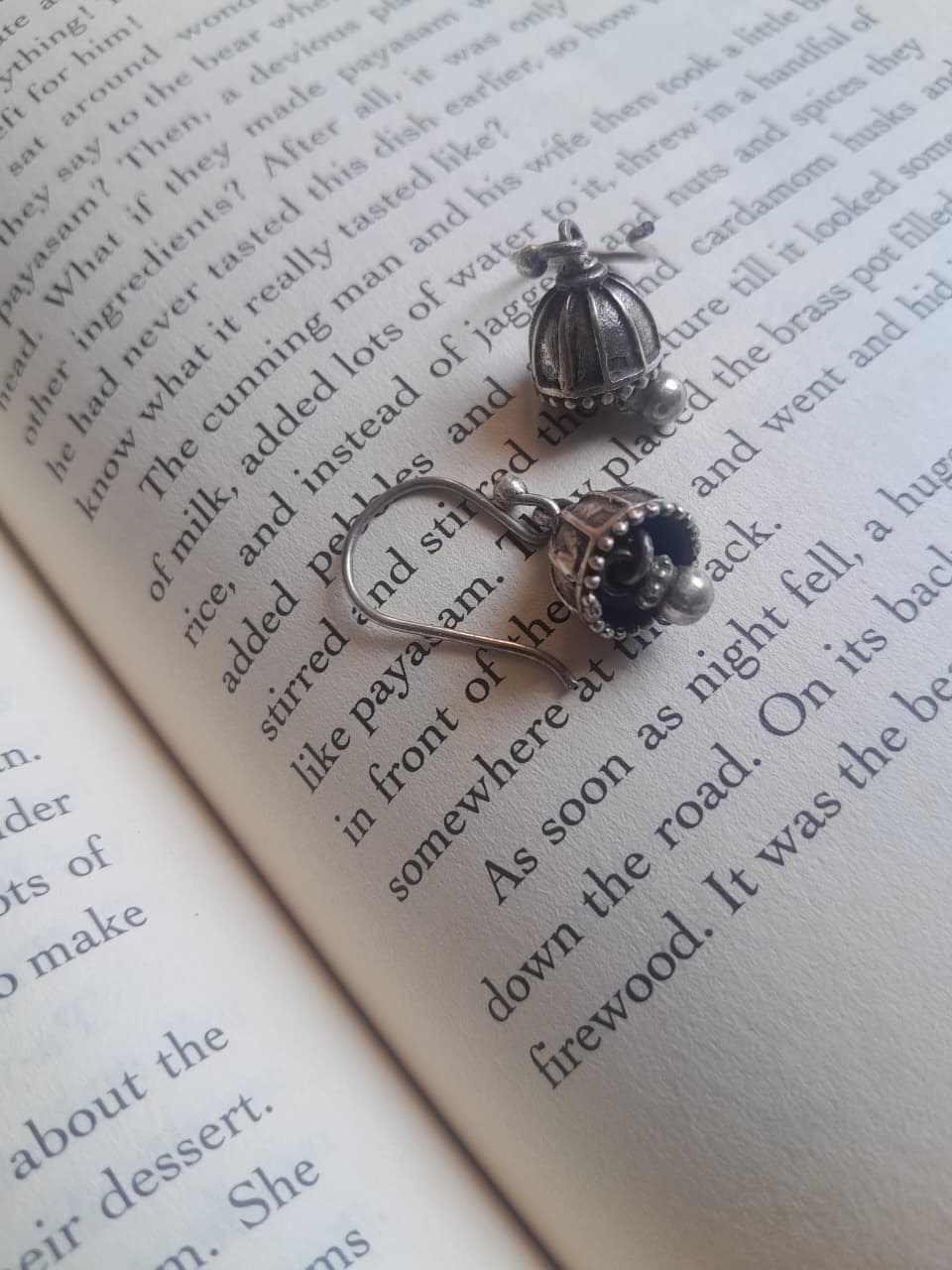 A pair of silver-toned jhumka earrings with decorative cabochon and floral details, displayed on the pages of an open book.