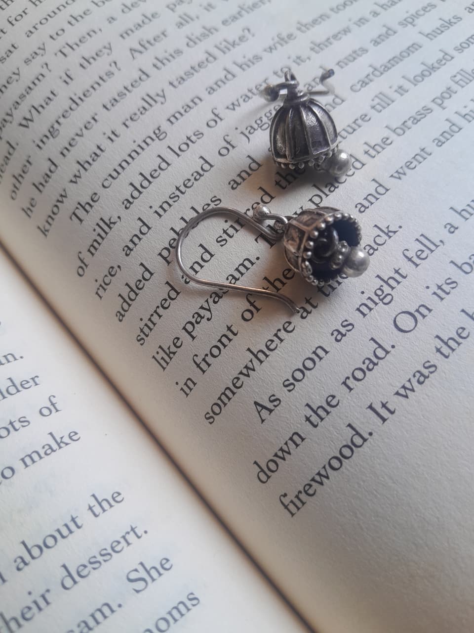 A pair of silver-toned dangling earrings with a small cage-like top and a dark floral charm, resting on open book pages with printed text.