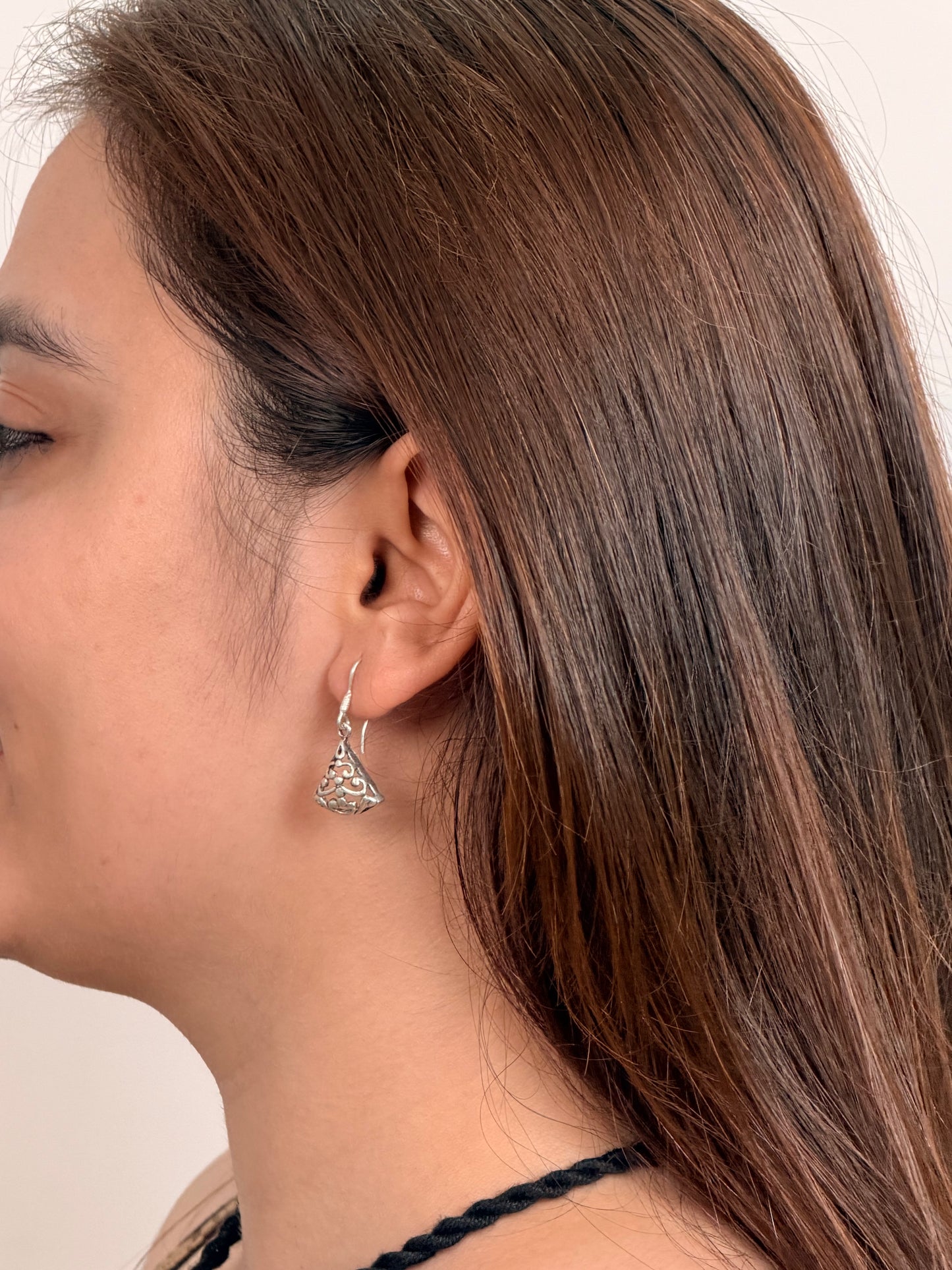 A close-up side profile of a woman with long brown hair wearing a small silver-toned triangular dangle earring with an intricate filigree pattern.