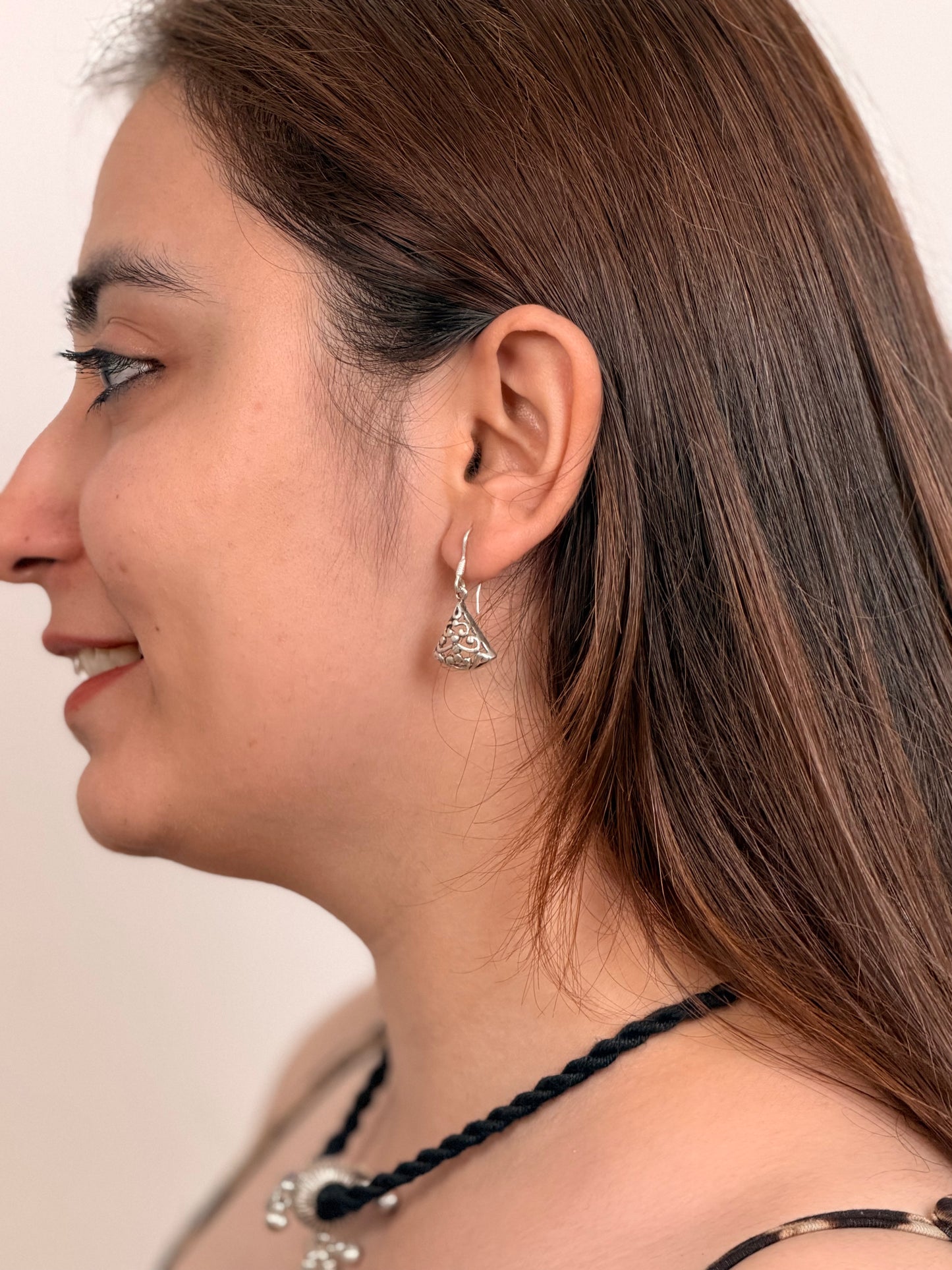 A close-up side profile of a woman with long brown hair wearing a silver triangular dangle earring with clear stones, along with a black braided necklace.
