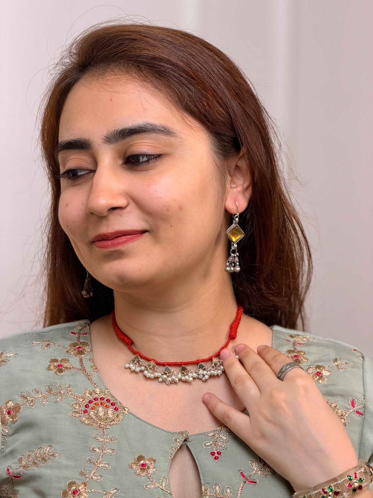 A woman wearing a red cord necklace with a silver-toned floral pendant, matching dangling earrings, and a ring, paired with a light sage green embroidered top.