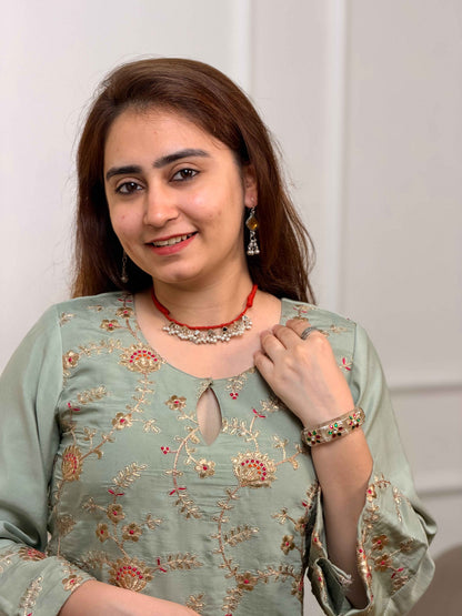 A woman with brown hair is wearing a sage green embroidered kurta with floral patterns, a red necklace, and a matching bracelet, set against a plain light gray background.