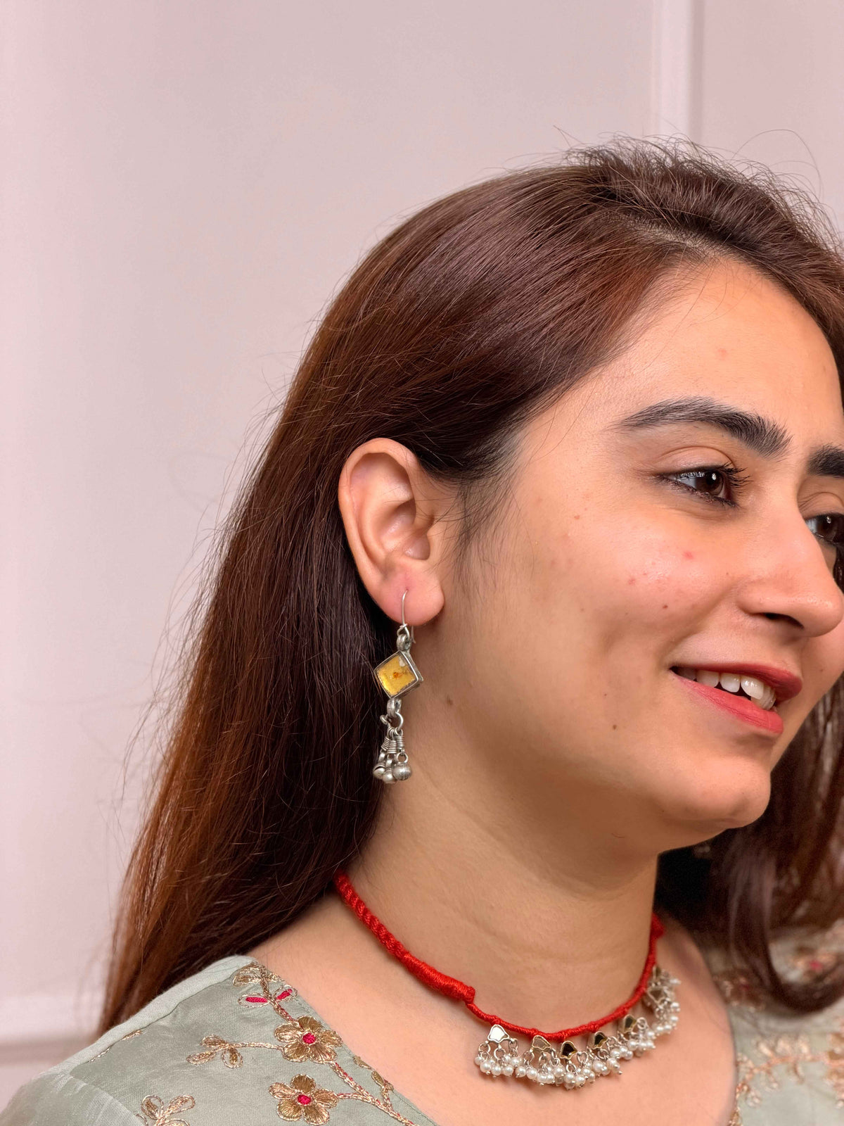 A close-up side profile of a woman with long brown hair, wearing a silver-toned traditional jhumka earring with a yellow stone and a red cord necklace with silver embellishments.
